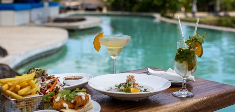 margarita and mojito cocktail and food served at a wooden table overlooking the pool